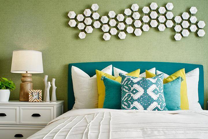 white hexagonal 3D wall art installed on sage green wallpapered wall, teal headboard, white bedding with white, yellow, and teal pillows, white bedside table, wood table lamp, potted plant, table accessories in bedroom, Indigo