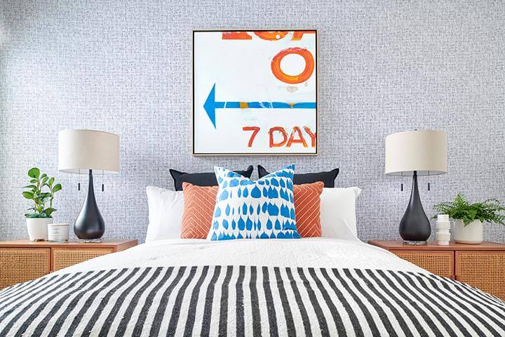 gray and white small patterned wallpaper, white bedding with orange and blue accent pillows, black and white striped blanket, wood and wicker bedside tables, black table lamps in bedroom, PGA West