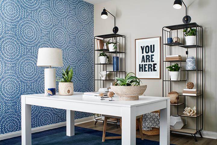 blue circular patterned wallpaper, white desk, two black metal bookshelves, white table lamp, greenery, blue area rug in home office, Bayberry